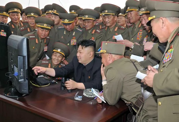 Kim Jong Un smiling and pointing at a computer monitor, surrounded by laughing uniformed military officers