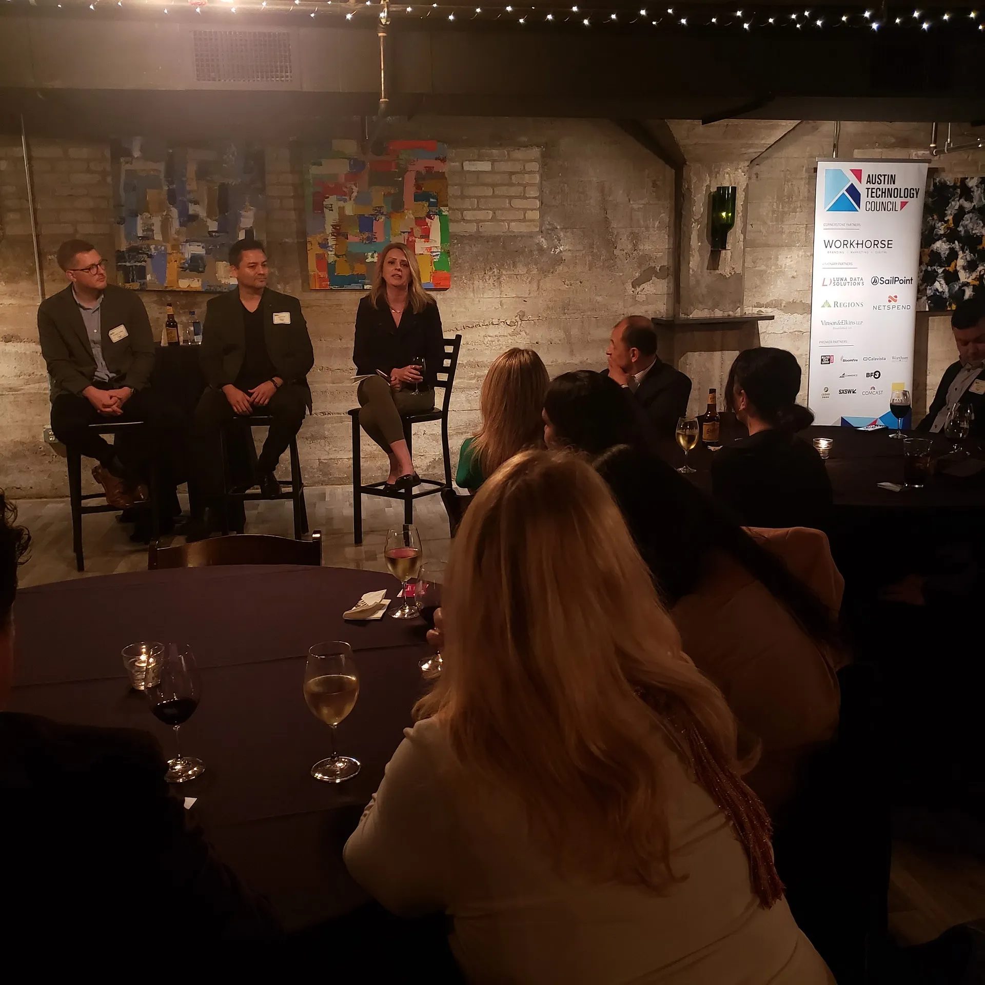 Three panelists on stools speaking to an audience seated at tables, beside an Austin Technology Council sponsor banner