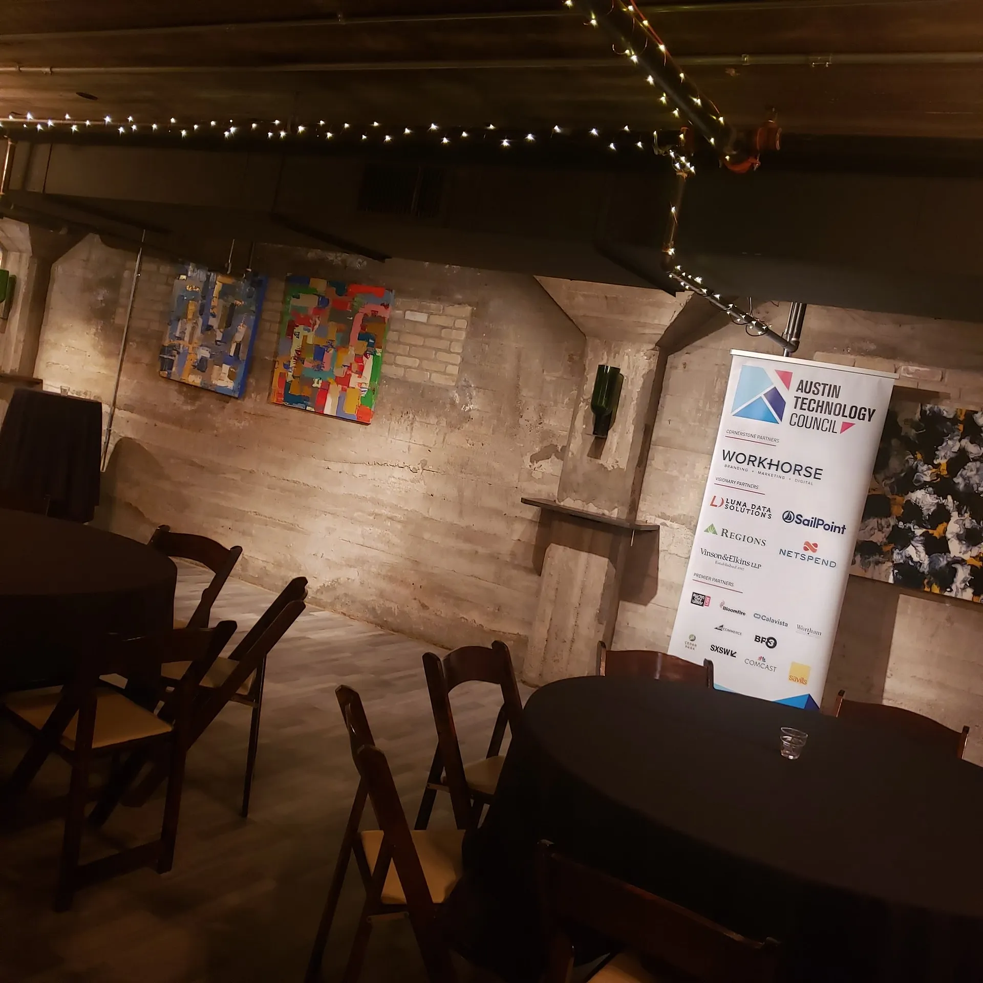 Empty event venue with string lights, round tables, colorful wall art and an Austin Technology Council sponsor banner