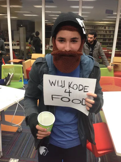 Teen in a knit beard beanie holding a handwritten "Will kode 4 food" sign and a paper cup in a library at night
