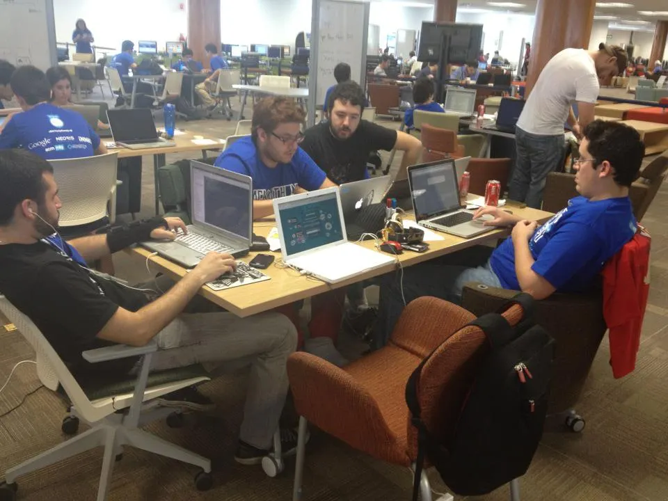 Icalia Labs' founders working on laptops at a shared table in a crowded hackathon space
