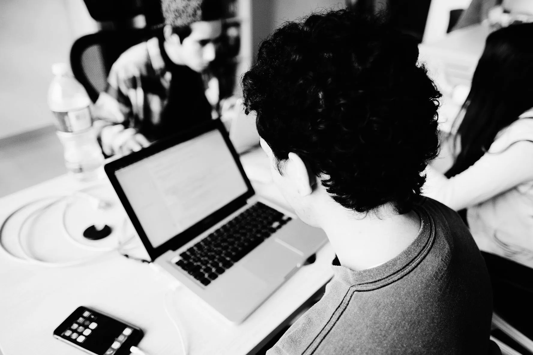 Black-and-white photo of a person with curly hair working on a laptop at a shared table with colleagues