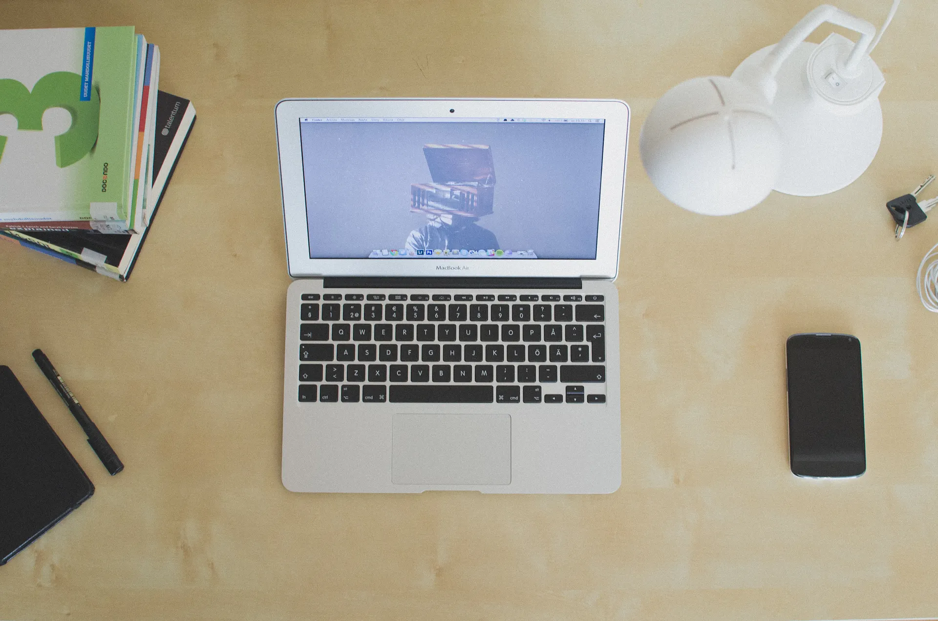 Overhead view of a laptop on a light wooden desk with a stack of books, a white lamp, keys, a pen, and a smartphone