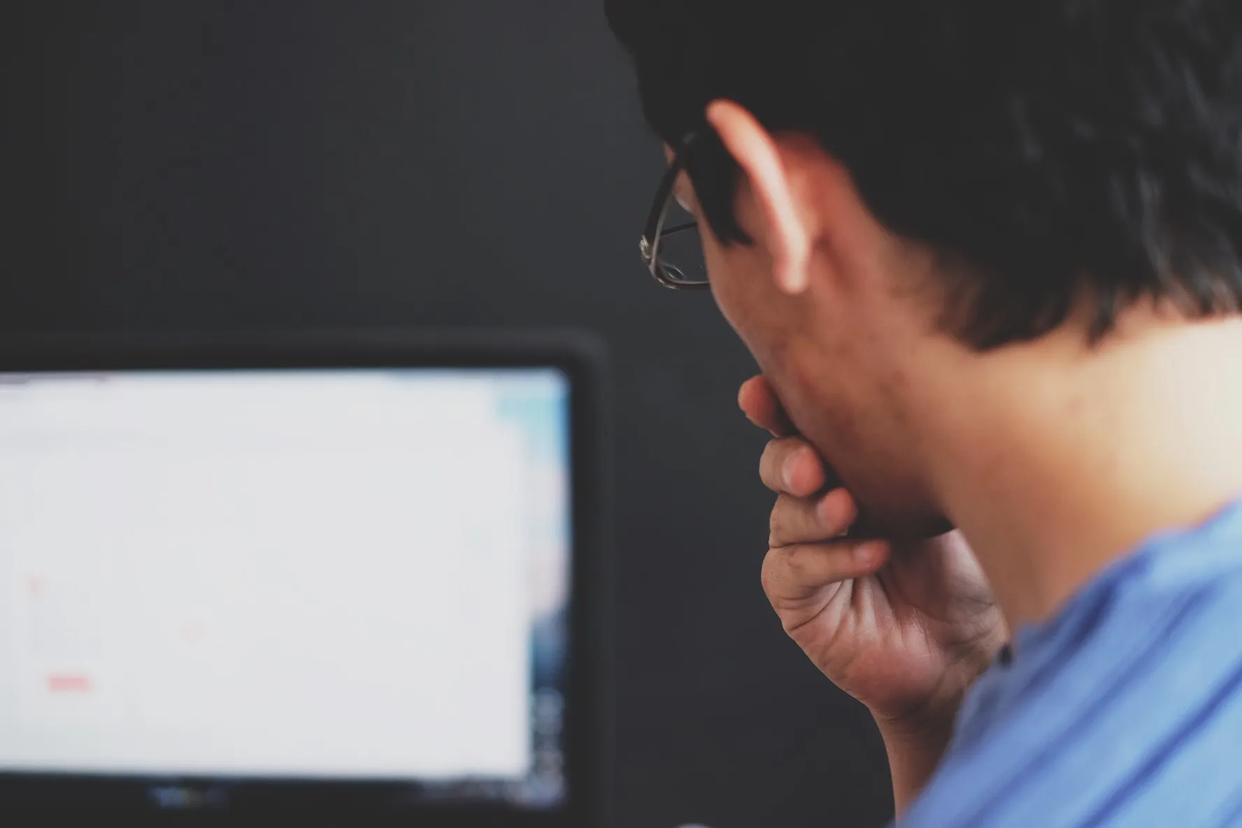Person in glasses resting their chin on one hand while looking at a blurred computer monitor